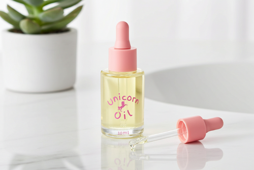 Bottle of 'Unicorn Oil' with a pink dropper on a white surface with a plant in the background