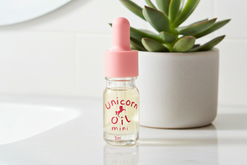 Bottle of 'Unicorn Oil mini' with pink cap on a white surface with a plant in the background