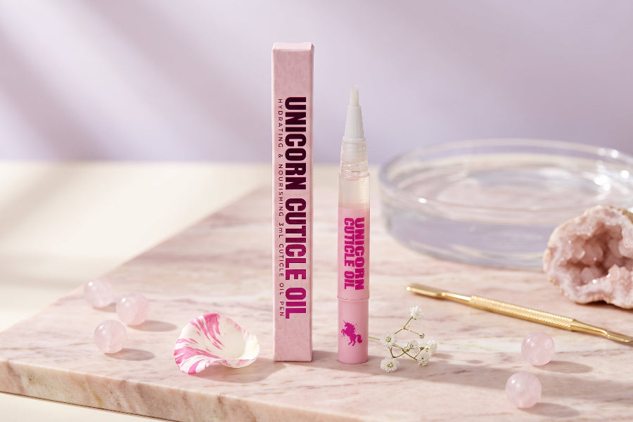 A pink cosmetic tube labeled 'UNICORN CUTICLE OIL' stands next to its matching pink box on a marble slab, styled with pink crystals, a rose petal, and a gold tool against a soft lavender background.