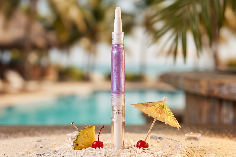 A single cuticle oil pen filled with shimmering lavender-purple liquid stands upright in a sandy beach setting with a blurred pool and palm trees in the background. Small props including a cocktail umbrella, pineapple wedge, and cherries are placed around the base.