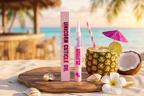 Unicorn Cuticle Oil treatment shown with its pink box and tube on a beach-themed board, surrounded by a pineapple drink, coconut, shells, and flowers.