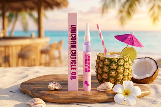Unicorn Cuticle Oil treatment shown with its pink box and tube on a beach-themed board, surrounded by a pineapple drink, coconut, shells, and flowers.