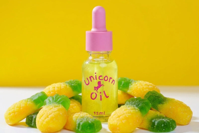 A small clear bottle labeled 'Unicorn Oil' with a pink dropper cap stands centered on a white surface, surrounded by yellow and green gummy candies against a bright yellow background.
