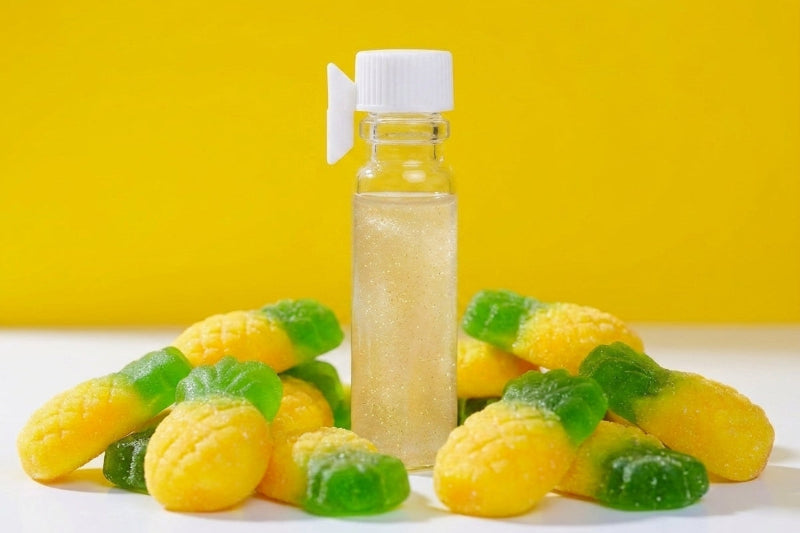 A small clear bottle filled with a pale yellow glittery liquid and topped with a white cap, surrounded by yellow-and-green gummy candies shaped like pineapples on a white surface against a bright yellow background.