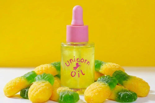 A small clear bottle of yellow 'Unicorn Oil' with a pink dropper cap, surrounded by yellow and green gummy candies on a white surface against a bright yellow background.