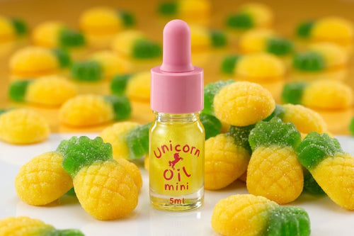 A small glass dropper bottle labeled 'Unicorn Oil mini 5ml' with a pink cap, surrounded by yellow and green pineapple-shaped gummy candies on a white surface with a warm yellow background.