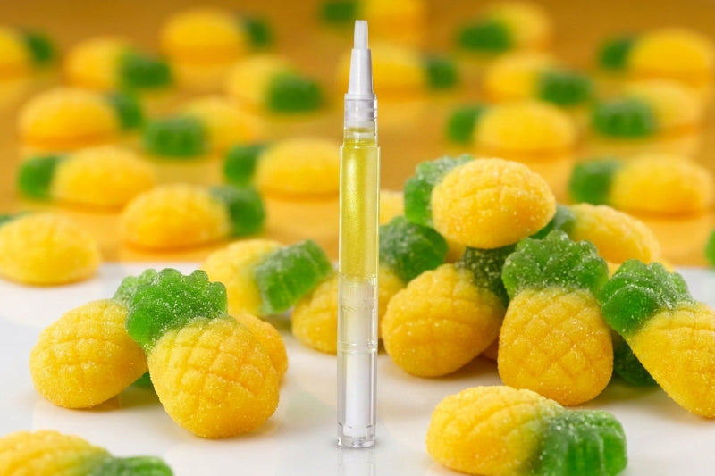 A clear glass dropper vial filled with pale yellow liquid stands upright in the center, surrounded by pineapple-shaped gummy candies in yellow and green on a white surface with a warm yellow background.