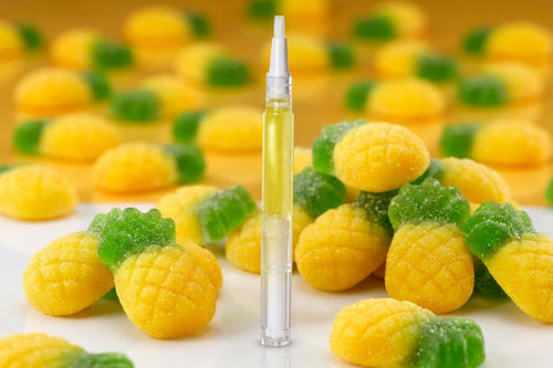 A clear glass dropper vial filled with pale yellow liquid stands upright in the center, surrounded by pineapple-shaped gummy candies in yellow and green on a white surface with a warm yellow background.