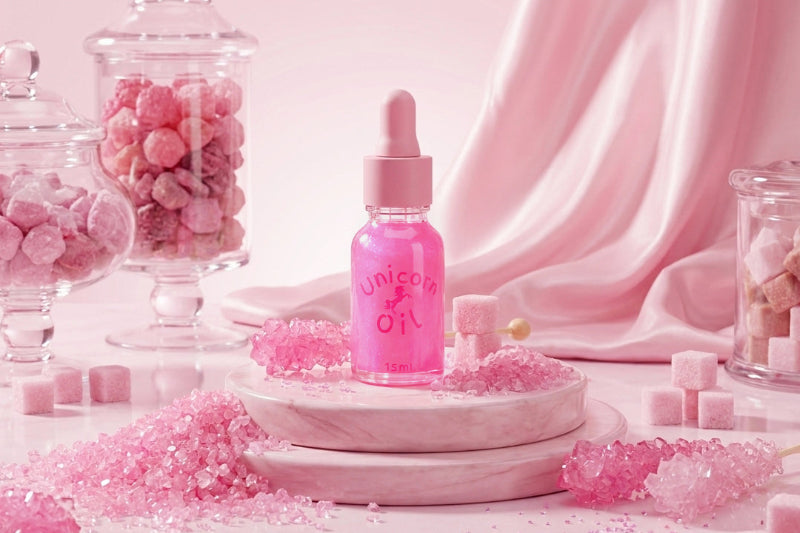 A pink dropper bottle labeled 'Unicorn Oil' sits on a marble pedestal surrounded by pink sugar crystals, pink candy cubes, and glass jars filled with pink candies against a draped pink satin backdrop.