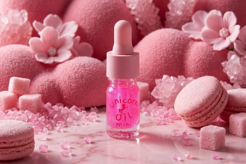 A small pink dropper bottle labeled 'Unicorn Oil Mini' stands centered on a pink marble surface, surrounded by pink macarons, pink sugar cubes, pink flower decorations, and pink textured spheres in the background.