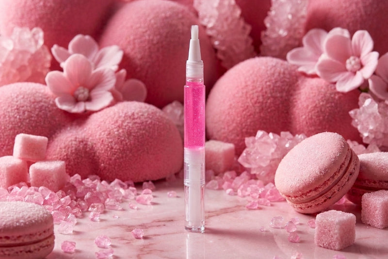A pink cosmetic dropper bottle stands upright on a pink marble surface, surrounded by pink macarons, sugar cubes, pink crystals, and soft pink spheres in the background.
