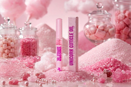 Pink cosmetic product packaging for 'Unicorn Cuticle Oil' stands upright on a pink surface surrounded by candy-themed props like jars of pink candies, cotton candy, and sugar crystals.