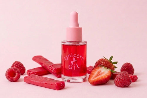 A small clear bottle of pink 'Unicorn Oil' with a pink dropper cap stands centered on a pale pink background, surrounded by red strawberry slices, whole raspberries, and several pink wax melts or solid bars.