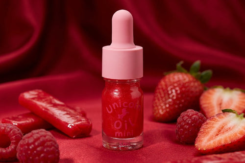 A small glass dropper bottle filled with bright red liquid, labeled 'unicorn oil mini,' centered on a deep red fabric background with strawberries, raspberries, and red candy pieces around it.