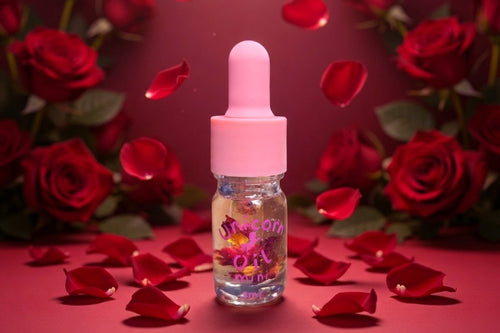A small clear bottle of rose oil with a pink dropper cap stands centered on a red surface surrounded by scattered rose petals and red roses in the background.