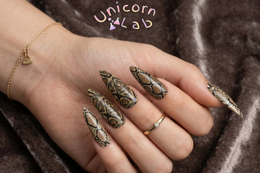 Hand with intricately designed nails on a brown fabric background, featuring the Unicorn Lab brand.