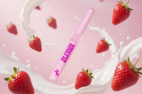 A pink cosmetic tube labeled 'UNICORN CREAM OIL' floating in a stylized scene with strawberries and white milk splash graphics on a pink background.