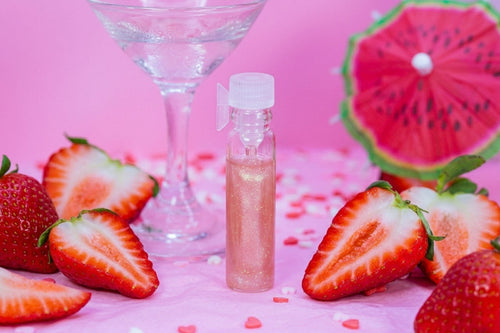 A small clear bottle filled with sparkling pink liquid sits on a pink surface surrounded by sliced strawberries and a watermelon slice prop, with a martini glass in the background.