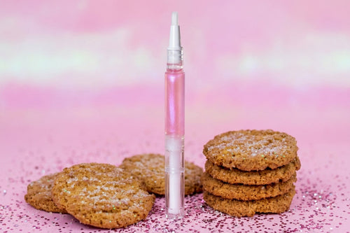A pink cuticle oil  in a clear tube with a white applicator tip, standing upright between several sugar-dusted cookies on a pink glittery background.