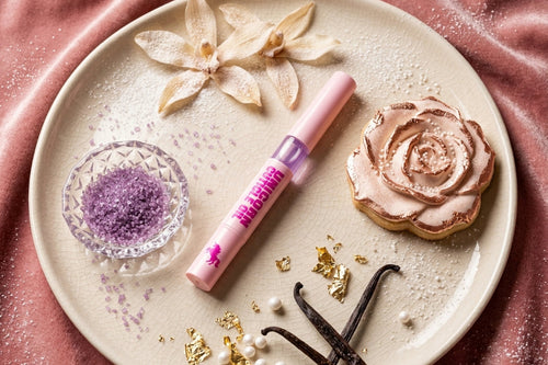 A pink cosmetic tube of cuticle oil lies on a round beige plate, styled with a pink rose-shaped cookie, purple sugar in a small glass dish, vanilla beans, gold leaf, and dried flowers on a pink satin background.