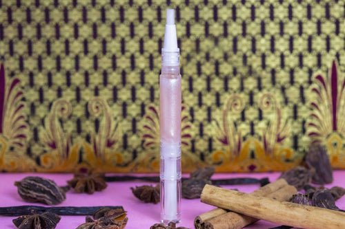 A clear plastic cosmetic tube with a white applicator nozzle stands upright on a pink surface, surrounded by cinnamon sticks, star anise, and vanilla beans in front of a patterned yellow and black fabric backdrop.