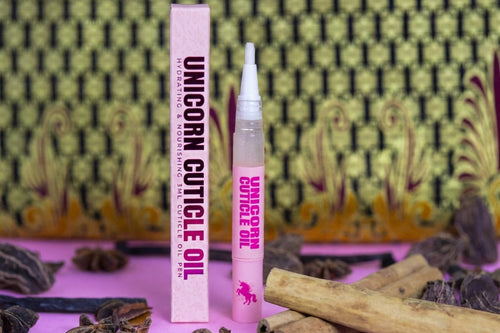 Unicorn Cuticle Oil set with a pink box and applicator tube, styled with cinnamon sticks and dried herbs on a pink surface against a patterned green and gold background.