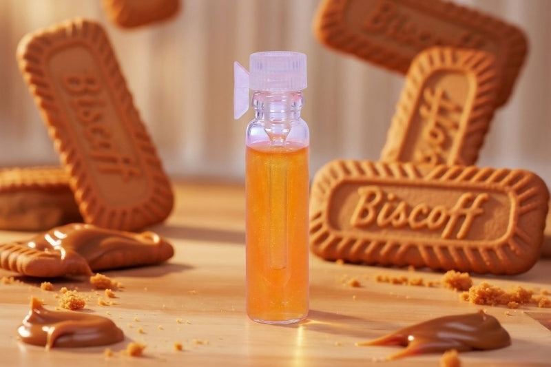 A small clear bottle filled with amber-orange liquid stands on a wooden surface, surrounded by Biscoff cookies, cookie crumbs, and caramel-like spread in a warm styled setting.