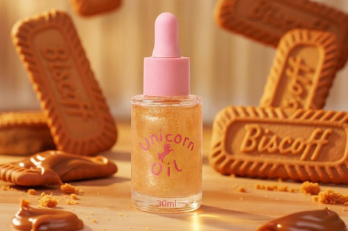 A 30ml bottle of Unicorn Oil serum with a pink dropper cap, filled with shimmering gold liquid. The bottle is styled on a wooden surface surrounded by Biscoff cookies, caramel drizzles, and crumbs, with more cookies floating in the background.