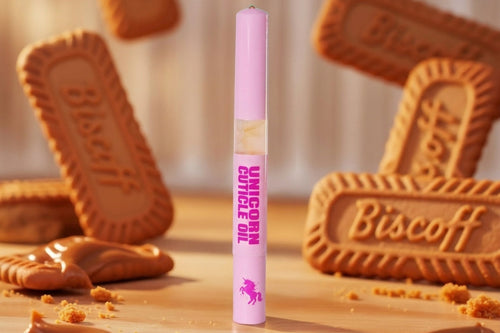 A pink cosmetic tube labeled 'Unicorn Cuff Oil' stands upright on a wooden surface, surrounded by Biscoff cookies and caramel drizzle in a warm, styled setting.