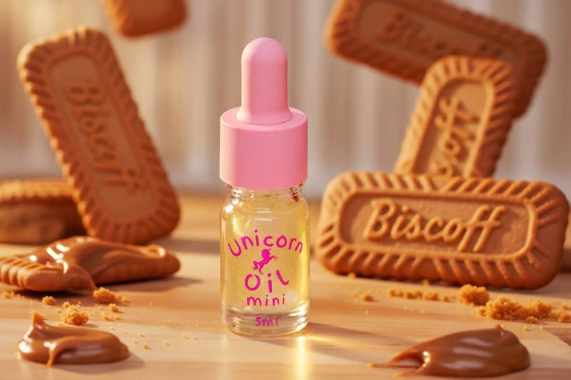 A small glass dropper bottle labeled 'Unicorn Oil Mini' with a pink cap stands on a tabletop surrounded by Biscoff cookies, cookie crumbs, and caramel drizzle in a warm, styled setting.