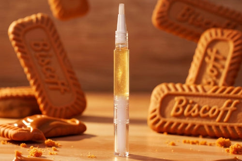 A clear cylindrical cosmetic tube filled with golden yellow liquid stands upright on a wooden surface, surrounded by Biscoff cookies and cookie crumbs in a warm, styled setting.
