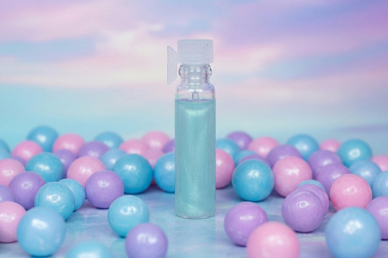 A small clear spray bottle filled with pale blue liquid stands upright in the center, surrounded by many pastel pink, blue, and purple glossy balls on a soft pastel surface with a matching sky-like background.