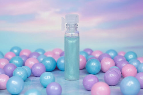A small clear spray bottle filled with pale blue liquid stands upright in the center, surrounded by many pastel pink, blue, and purple glossy balls on a soft pastel surface with a matching sky-like background.