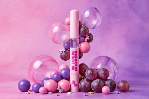 A pink cosmetic tube labeled 'UNICORN  OIL' stands upright in a styled studio setup with pink and purple balloons, glossy spheres, and a few grapes against a pink-purple gradient background.