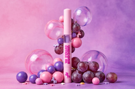 A pink cosmetic tube labeled 'UNICORN  OIL' stands upright in a styled studio setup with pink and purple balloons, glossy spheres, and a few grapes against a pink-purple gradient background.