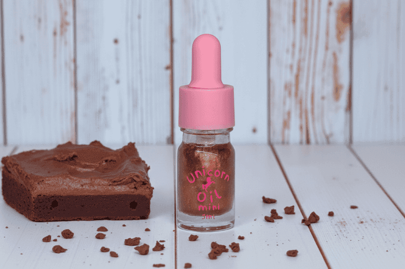 Small bottle labeled 'Unicorn Oil Mini' with a pink dropper next to a piece of chocolate cake on a wooden surface.