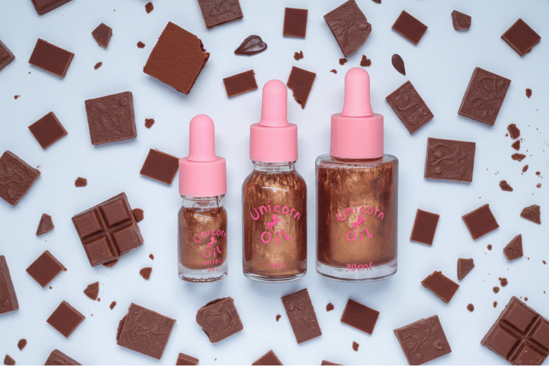 30ml glass dropper bottle of Chocolate Brownie Unicorn Cuticle Oil featuring golden shimmering oil and a professional label. with broken chocolate pieces