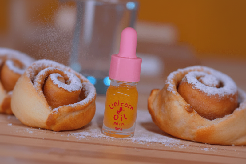 5ml glass dropper bottle of Cinnamon Scroll Unicorn Cuticle Oil showing a golden spiced oil with swirling shimmer in a bakery with cinnamon scrolls. 