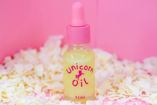 A 15ml glass dropper bottle of 'Unicorn Oil' filled with pale yellow liquid, set against a solid pink background and surrounded by white flower petals.