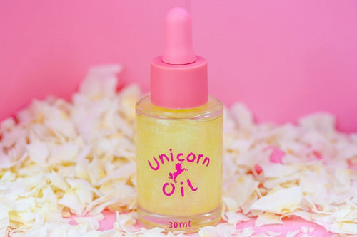 A 30ml bottle of 'Unicorn Oil' with a pink dropper cap, filled with yellow oil and decorated with a pink unicorn logo, set against a pink background with scattered white flower petals.