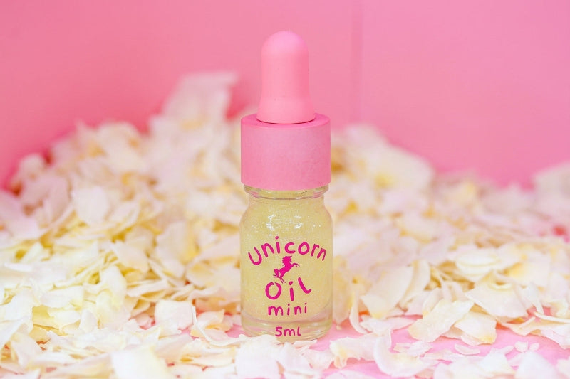 A 5ml dropper bottle of Unicorn Oil Mini with pale yellow oil inside, set against a pink background and surrounded by scattered white flower petals.