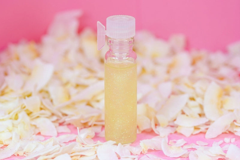 A small clear plastic bottle filled with a pale yellow glittery liquid, standing upright on a pink surface surrounded by scattered white flower petals.