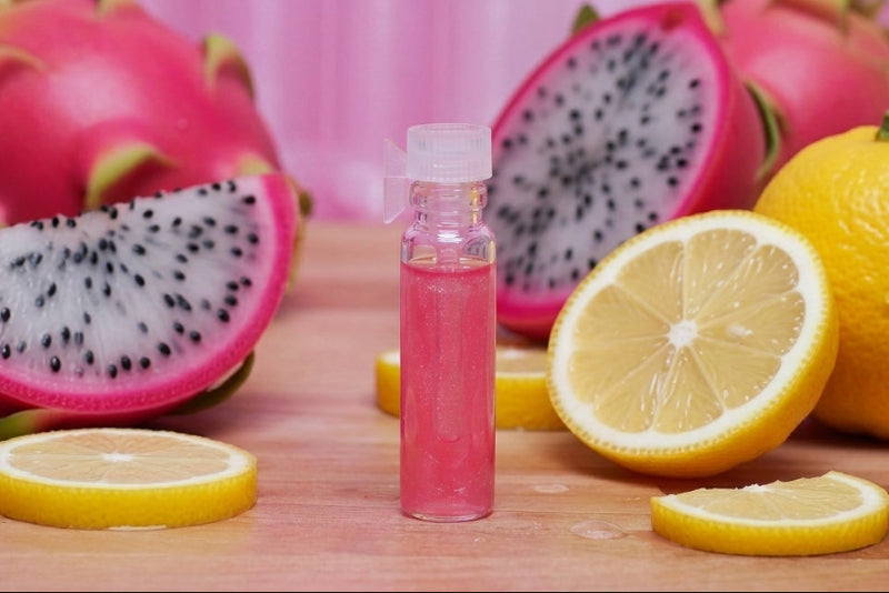 A small clear bottle filled with pink liquid stands on a wooden surface, surrounded by sliced dragon fruit and lemon halves, with a pink striped backdrop behind.