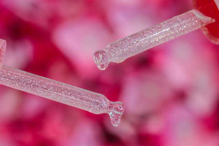 Close-up of two nail serum droppers with sparkly cuticle oil dripping with a pink blurred background