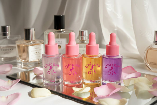 Four bottles of 'Unicorn Oil' with pink droppers on a reflective surface with flowers.