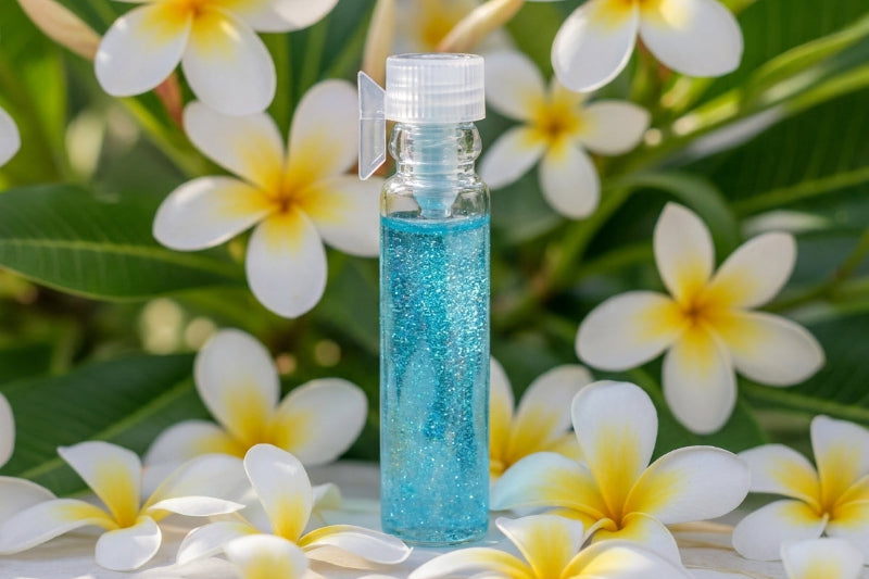 A small clear bottle filled with sparkling light blue liquid and topped with a white cap, placed among white and yellow plumeria flowers with green leaves in the background.