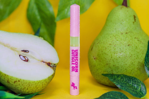 A pink tube labeled 'UNICORN CUTICLE OIL' stands upright on a bright yellow surface, flanked by green pears and leaves. The product packaging features a pink cap and base with a light green band in the middle.