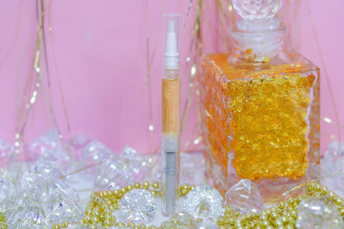 A clear cosmetic tube filled with shimmering gold liquid stands beside a large clear glass bottle packed with gold flakes, set against a pink background with decorative beads and crystal-like props.