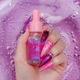Hand with pink and purple glittery nails holding a small bottle labeled 'Unicorns Oil' against a purple bubble. Grape Soda Unicorn 15ml Cuticle Oil. A bottle of scented shimmer nail serum in the scent grape soda. Part of the sweet  candy scented cuticle oil range at Unicorn Lab. background.