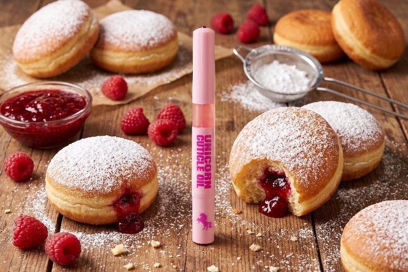 A pink tube labeled 'UNICORN OIL' stands upright on a rustic wooden table surrounded by powdered donuts, fresh raspberries, and a small bowl of red jam.